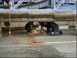 This photo shows three workers crawling under a structure. There are several orange cords running down to them.