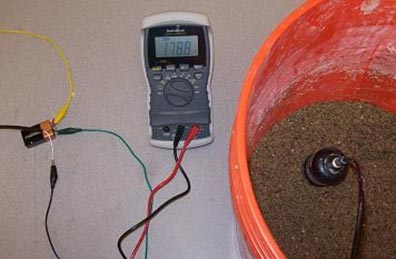 This photo shows a testing setup for a moisture cell. There is a red bucket containing sand, and the moisture gauge is sticking into the sand. A black cord is running for the moisture gauge to an apparatus that is giving a reading of 1,788. The apparatus also has a cord running to a battery that is connected to three other wires.