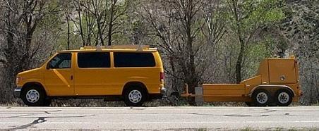 This photo shows a falling weight deflectometer (FWD), which is housed in a yellow trailer that has two axles. The FWD trailer is pulled by a yellow van. 