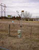 This photo shows mini site-specific seasonal monitoring program (SMP) weather station. It consists of a light green mechanical box as the base. In the middle of the base is a black rectangle with a yellow line across the middle. There is a pole extending upward from the base. Just above the base on the pole is a brown box. Near the top of the pole, there is an L-shaped pole attached to the left. At the top of the pole and the L-shaped pole, there are additional sensors. There are two brown stakes on either side of the mini SMP weather station. 