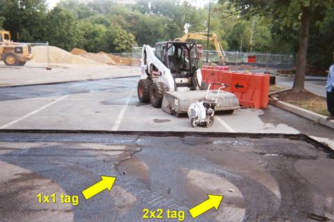 This photo shows parking lots where the tags are being embedded. In the background is the undisturbed pavement. Equipment, such as a saw and a small pavement brush are located on this pavement. In the foreground, the top layer of pavement has been removed. Two tags are placed in the middle of this pavement. An arrow points to the tag on the left labeled 1x1 tag. Another arrow points to the tag on the right labeled 2x2 tag.