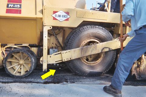 This photo shows a close-up of the paver as it is about to cover the tag with a layer of asphalt. An arrow points to the tag located between the lead tire and main tire.