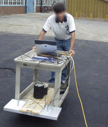 This photo shows a picture of a cart. The cart consists of a lower and top shelf. There are handles on the top shelf. Although they cannot be seen, it appears the cart is on wheels. The top of the cart holds a laptop and several wires connecting to the antenna, which is connected to the lower shelf. The antenna is connected to the lower shelf using wood. The operator is standing behind the cart.