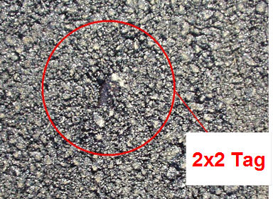 This photo indicates where a radio frequency identification tag has surfaced, which is represented by a larger void and discoloration in the pavement. The surfaced tag is circled and labeled 2x2 Tag.