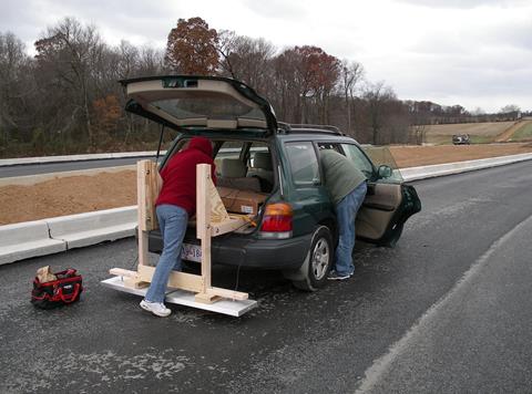 This photo depicts the antennae mounted from the back hatch of a station-wagon. The mount is made of 2-by-4 wood pieces that extend from the trunk out and connect to vertical posts that attach to the antennae that is mounted just above the pavement. The antennas consist of two rectangular metal pieces. Two operators are visible but they are working inside the car and details cannot be provided.