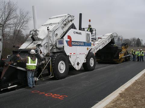 This photo shows the RoadTec® paver paving the upper lift of asphalt at Hampstead Bypass. The photo shows the paver in the background (right-hand side) where several technicians are working. The Roadtec® material transfer vehicle is feeding the asphalt mixture to the paver (middle), which is being filled by a dump truck (left side of picture), which is not visible. There is a technician between the dump truck and the material transfer vehicle.