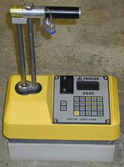 This photo shows the nuclear density gauge. At the bottom of the photo is a rectangular base that sits on the ground. From the base, another rectangular layer connects to the rectangular panel of the nuclear gauge. The panel consists of a keypad and a small screen that is located on the right side. On the left side, a rod with two shafts extends upward from the panel and has a handle at the top.