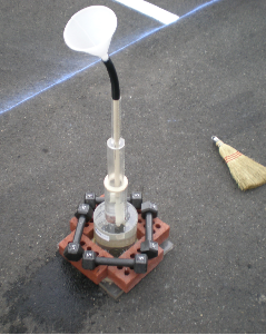 This photo shows the permeability test setup. The test is set up on the asphalt pavement. Directly touching the pavement is a clear plastic rectangle that has a large diameter cylinder connected to it at the center. There are four bricks placed on each side of this rectangle. Four 5-lb dumbbells are placed diagonally from the center of a brick to the center of another brick. Extending upward from the top of the first cylinder is a second cylinder of smaller diameter. Extending from this second cylinder is a third cylinder of yet even smaller diameter. Extending from the top of this third cylinder is a semi-flexible pipe that then connects to the bottom of a funnel. There is a broom head in the right-hand background of the photo.
