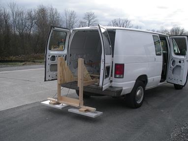This photo depicts the antennae mounted from the back of a cargo van that has the two rear doors open. The mount is made of 2-by-4 wood pieces that extend from the floor of the van out and connect to vertical posts that attach to the antennae that are mounted just above the pavement. The antennas consist of two rectangular metal pieces.
