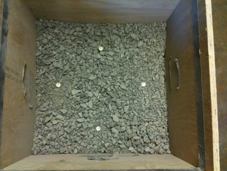 This photo shows a wooden box containing course aggregate from the top view. There are four tags placed within the coarse aggregate. The tags are located at the center of each of the four sides of the box, a distance of 4 inches (100 mm) away from the sides. The tags form a diamond located at roughly the center of the box. The tags are easily identified as circular and a different material than the coarse aggregate.