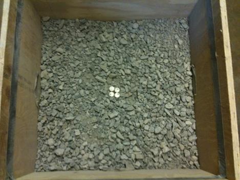 This photo shows a photo of a wooden box containing coarse aggregate from the top view. There are four tags placed within the coarse aggregate. The tags are located at the center of each of the four sides of the box, a distance of 12 inches (300 mm) away from the sides, which places them roughly in the center of the box and touching. The tags are easily identified as circular and a different material than the coarse aggregate.