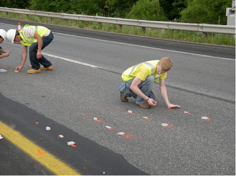 This photo shows the placement of a cluster of four groups of surface acoustic wave radio frequency identification tags in the foreground. Each group consists of placing four tags as illustrated in figures 97 and 98. The location for the placement of the tags was indicated by applying spray paint on the surface of the pavement. The location of the tags can be identified by circles, which are lighter in color than the pavement. A technician is working on one of the groups. In the background, two other technicians are working on another cluster of groups, but details cannot be seen.