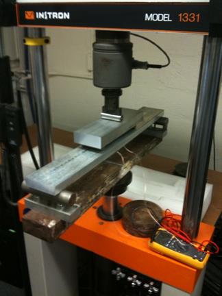 This photo shows the setup for testing the thin film in the machine using the four-point bending test as described in figure 140. In the photo, the four-point bending test setup is placed in the loading machine. The loading machine consists of a load cell at the center top of the loading machine and a base on which the test rests. Wires connected to the testing apparatus connect to an ohmmeter. The testing machine is an Instron® Model 1331.