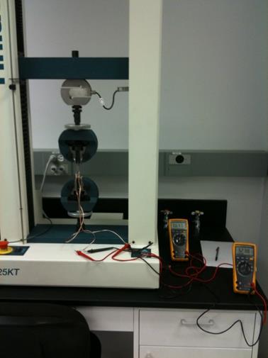 This photo shows the setup of the direct tension test using the dogbone specimen. The dogbone specimen is placed in the loading machine vertically. The machine consists of a load cell at the top that applies the displacement. Wires extend from the dogbone specimen and are connected to the ohmmeters.