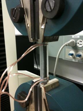 This photo shows a close-up of the direct tension specimen in the testing machine. The specimen is located between clamps in the center of the photo. Two wires extend from both the top and bottom of the specimen.