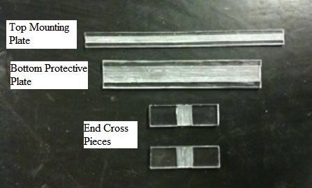This photo shows the four parts used for encapsulating the radio frequency identification (RFID) tag. The four parts are placed in a vertical line. The part on the top is labeled Top Mounting Plate and is 12 inches long by 0.4 inches wide and 0.095 inches thick polycarbonate. A longitudinal groove deep enough to accommodate the RFID tag is cut into the mounting plate. The next part is labeled Bottom Protective Plate and is 10.8 inches long and appears about twice as wide as the top mounting plate and is also made of polycarbonate. A longitudinal groove deep enough to fit the RFID tag is cut into the mounting plate. The next two parts are labeled End Cross Plates and are 0.6- by 0.6-inch cross pieces of 0.18-inch thick polycarbonate. A vertical groove deep enough to accommodate the RFID tag is cut into the center of the cross pieces.