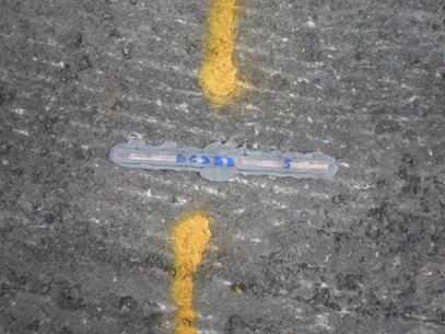 This photo shows the placement of the encapsulated tag on the roadway. The tag is placed in the horizontal direction. A notation on the tag appears to be a label but it is not legible. Two vertical lines are spray painted from the center of the tag, above and below the tag. The tag is 1 of the 16 that were sandwiched between two 0.4-inch wide polycarbonate plates.