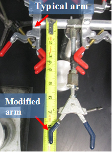 Typical arm and modified arm for the wrist action shaker. View of a typical wrist action shaker with several arms used in the foam index text for agitating mixtures containing air-entraining agent solution, fly ash and cement. A red arrow at the top indicates the typical arm. A red arrow at the bottom points to the modified arm. A ruler placed between these arms measures the distance between them.