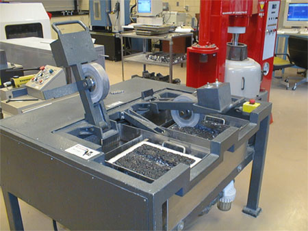 Figure 1. Photo. Hamburg Wheel-
Tracking Device without water. The machine has a heavy steel wheel that travels back and forth over a slab of asphalt mixture. The slabs are lifted in and out of the chamber, which is filled with water. The water controls the test temperature, so there is no environmental chamber.