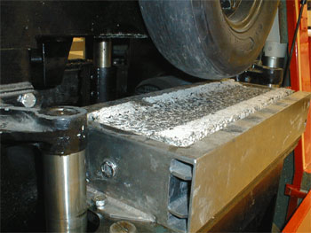 Figure 18. Photo. French Pavement Rutting Tester wheel and slab. This photo of the French Pavement Rutting Tester is taken very close to the tire. It includes a slab that has been tested. The slab has a rut in it from testing. Upward heaving of the mixture has occurred outside the wheelpath. The slab is held in place by a steel mold.