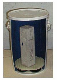 Figure 10. Photo. Concrete prism test—prisms stored over water in sealed containers. This photo of the A S T M C1293 concrete prism test shows a rectangular concrete sample standing inside a plastic bucket. Test procedures are described in section 3.4.2 of the text.