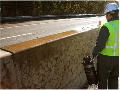 Figure 21. Photo. Spraying 30-percent lithium nitrate solution with handheld spray applicator on a barrier wall near Leominster, Massachusetts. This photo shows a worker using a handheld spray applicator to topically spray lithium nitrate on an A S R affected concrete barrier. The barrier is part of a median structure on a highway. There is extensive cracking in the barrier, and the top of the barrier is moistened with the lithium nitrate.