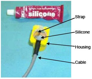 Photograph shows a housing filled with electronic grade silicone sealant/adhesive. A tube of silicone adhesive also appears in the photo. Arrows indicate the locations of the applied silicone, cable, strap, and housing.