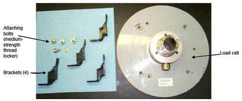 Photograph shows a load plate with mounting brackets. Arrows indicate the locations of the attaching bolts open parenthesis use medium-strength thread locker close parenthesis, four brackets, and load cell.