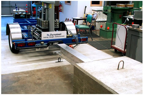 Figure 10. Photo. FWD calibration beam and inertial block. This photo depicts a Dynatest<sup>®</sup> falling weight deflectometer (FWD) positioned in front of the old beam and block system developed for the 1994 Strategic Highway Research Program protocol. The FWD is positioned in front of a large aluminum beam attached to a large concrete block in a laboratory. It is sitting on a concrete slab isolated from the rest of the concrete floor. A sensor holder is attached to the isolated concrete slab just below the front end of the aluminum beam.