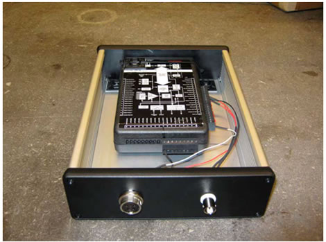 Figure 11. Photo. Box for DAQ wiring protection. The photo shows a KUSB data acquisition board (DAQ) inside an uncovered box. Wires from the DAQ are attached to two connectors on the front end of the box. The box is aluminum on the left, bottom, and right side. The front and back end of the box are made of hard plastic.