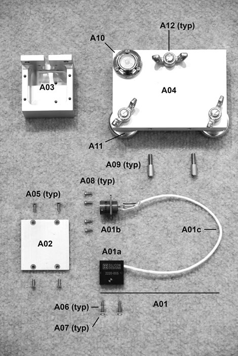 Figure 114. Photo. Parts for accelerometer box assembly. The photo shows an enlarged view of the accelerometer box, and the individual parts are numbered. The upper left shows the rectangular shell of the accelerometer box assembly. The upper right shows the calibration platter with bubble level in the upper left corner and wing nuts attached to leveling feet in three locations. Two knurled nuts are aligned with screw holes on the platter. The lower right shows the electrical connector and accelerometer with connecting wiring. The lower left shows the bottom plate with four machine screws.