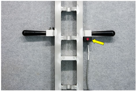 Figure 118. Photo. Calibration stand with handles, bubble level, and pushbutton. This photo shows a view of the placement of the calibration stand handles, bubble level, and pushbutton. The stand is vertical on a flat surface with two handles coming out of each side of it. The pushbutton is attached to the calibration stand using Velcro ® below the handles. A yellow arrow points to the pushbutton on the right side of the stand.