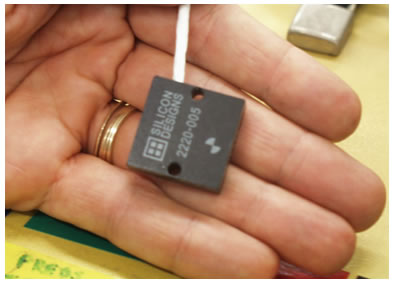Figure 12. Photo. Silicon Designs model 2220 accelerometer. The Silicon Designs model 2220 accelerometer is shown in a researcher’s hand. The accelerometer is in a thin black 0.975-inch (25-mm)-wide, square-shaped case with two small holes on the left and right sides used to attach the accelerometer inside the accelerometer box. A diameter target symbol that is smaller than 0.195 inches (5 mm) on the right side of the upper surface shows the actual location of the accelerometer in the case.