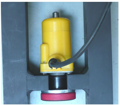 Figure 37. Photo. Attachment of a Carl Bro geophone in the stand. This photo shows a close-up of an attached Carl Bro geophone in the calibration stand. The components are the Carl Bro geophone, which is a yellow plastic cylinder with a metal plate on the bottom, a wire out the front, a hidden bolt out the bottom, the geophone stand with a notched shelf for placing the geophone, a large lower washer, and a black knurled knob.