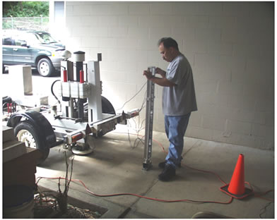 Figure 38. Photo. On-site FWD calibration at Hawaii Department of Transportation Materials Lab. This photo shows a Hawaiian Dynatest® falling weight deflectometer (FWD) in a breezeway at the Hawaii Department of Transportation Materials Lab. FWD is on the left side of the photo with its back facing the right. An employee is holding the geophone sensor stand with geophones attached during a calibration trial. The stand is bolted to the concrete floor 
