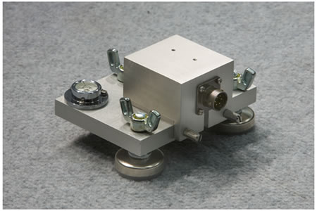 Figure 47. Photo. Accelerometer box. This photo shows a perspective view of an aluminum accelerometer box attached to an aluminum calibration platter. The accelerometer box is attached to the front of the platter by two knurled thumb screws. Three wing nuts attached to plastic feet are used to level the platter. A bubble level on the back left corner of the platter is used to confirm that the platter is level.