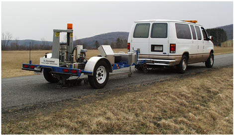 Figure 6. Photo. Dynatest ®  model 8000 trailer-mounted FWD. This photo shows the Dynatest ® model 8000 trailer-mounted falling weight deflectometer (FWD) attached to a white tow vehicle on a rural road with landscape on both sides of the road. The load plate of the FWD is in the down position ready for testing.