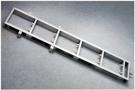 Figure 68. Photo. Seismometer calibration stand. This photo shows a seismometer calibration stand. The stand, which is composed of aluminum, is positioned diagonally on a flat surface, but it is normally used in a vertical position. It is a long rectangle with bars at each end going between the two vertical channel pieces. Five shelves are positioned between the channels to accommodate the geophones and accelerometer. Four small aluminum blocks are positioned 
near the top to accommodate handles and a bubble level.
