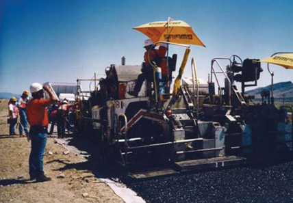 Large asphalt paving equipment sitting at edge of road in bright sun, with construction crews standing by next to the vehicles and on board the vehicles.