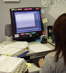 Woman seated at office desk with computer monitor and keyboard in front of her and stacks of paper next to her.