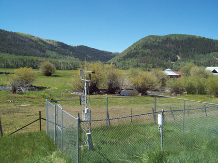 A rural scene with a weather station enclosed in chain link fencing in the foreground. A rain collector is positioned on the fence. Trees and a few buildings appear in the background and mountains further distant.