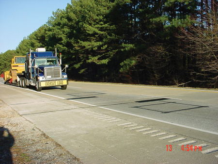 A stretch of highway with weigh-in-motion equipment embedded in the pavement is shown with a tractor trailer approaching. Trees are growing at the side of the highway.