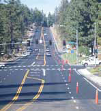 A four-lane stretch of highway is shown with asphalt pavement and work zone cones closing off the right lane. An intersection with traffic lights and cross walks appears in the middle distance, with light traffic further away.