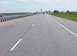Two lanes of a four-lane asphalt paved highway are shown with a guard rail in the median strip. There is a white dotted line between the lanes and a solid line separating the right lane from an asphalt shoulder.