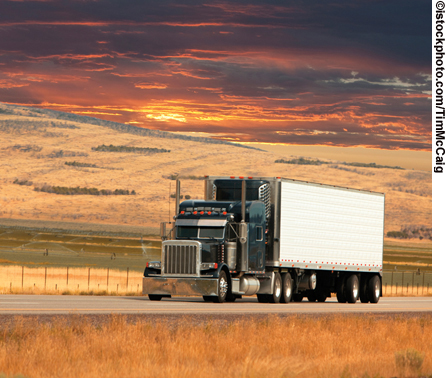 A tractor trailer is shown on a highway in hilly, rural countryside with the sun setting in the background.
