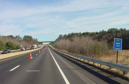 A stretch of four-lane highway is shown with a concrete barrier in the median strip. Traffic is moving on the far side of the median. The pavement is asphalt. On the near side, orange work zone cones are standing on the white dotted line between the two lanes, a solid white line separates the right lane and the asphalt shoulder, which contains a noise strip, and a construction vehicle and another vehicle are visible in the distance. At the side of the road is a blue sign reading "Road Test 331001."