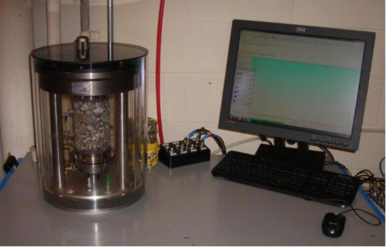 Figure 1. Photograph. Dynamic Modulus Test Setup in the Asphalt Mixture Performance Tester. This photograph shows an asphalt sample in the Asphalt Mixture Performance Tester apparatus. The apparatus is connected to a computer and monitor, which displays the dynamic modulus testing setup.