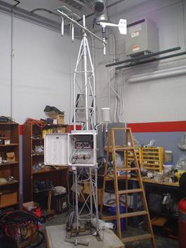 Figure 6.3. Photo. Prototype weather station used by LTPP regional support contractors for in-house training of staff and to calibrate equipment components. A weather station, tall frame with white case attached about 4 ft from the base, stands in a workshop. A stepladder stands beside it. The door of the case is open and equipment can be seen inside.