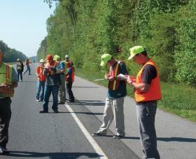 Figure 6.13. Photo. Workers in safety helmets and vests holding clipboards and examining pavement from roadside.