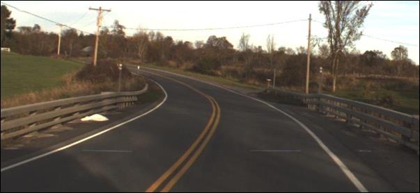 This photo shows the approach to a geosynthetic reinforced soil integrated bridge system (GRS-IBS) bridge in the eastbound direction. This bridge is a two-way one-lane road and is on a curve that turns to the left and is located on County Road 12 over Malterna Creek in Lawrence County, NY.